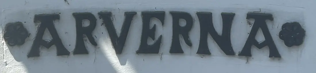 Photo d'une plaque de nom de maison : le mot ARVERNA en lettres pleines à empattements, encadré de deux rosaces, sur un mur clair strié d'ombres de feuillage.